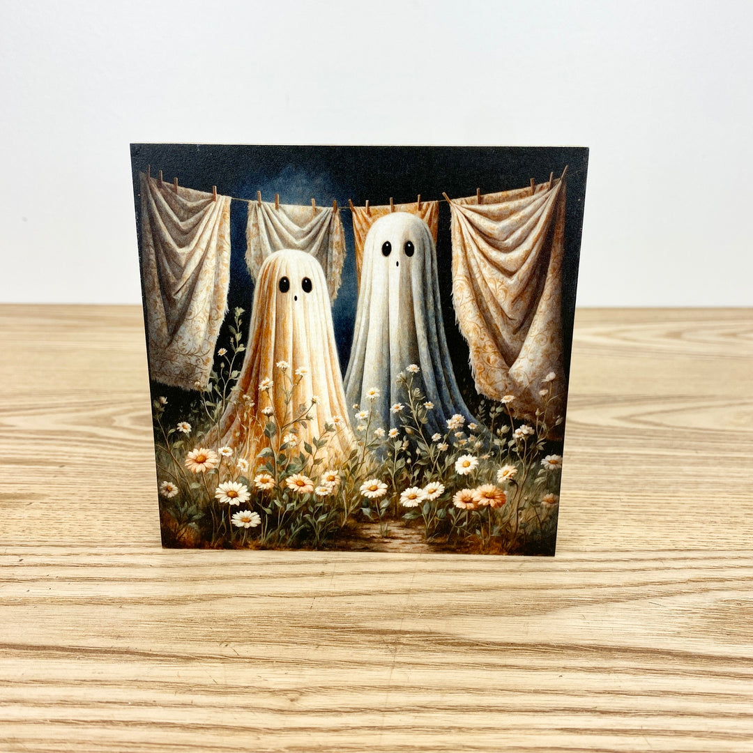 Two ghostly figures hanging on a line with curtains and flowers in the background on a wooden surface.