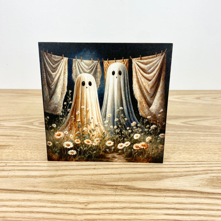 Two ghostly figures hanging on a line with curtains and flowers in the background on a wooden surface.