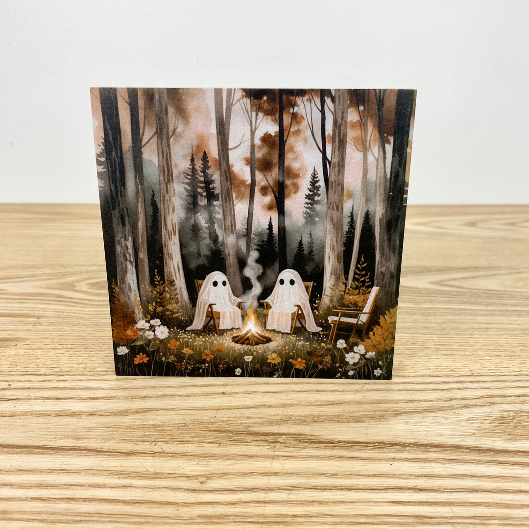 Card with two ghosts by a campfire in a forest setting on a wooden surface.