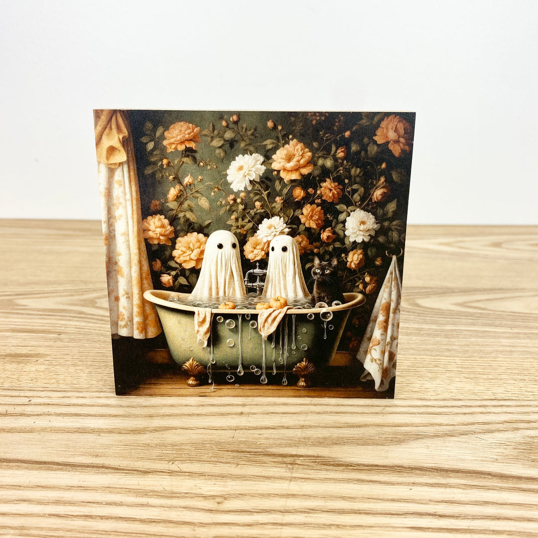 Decorative card with ghosts in a bathtub and floral background on a wooden surface.