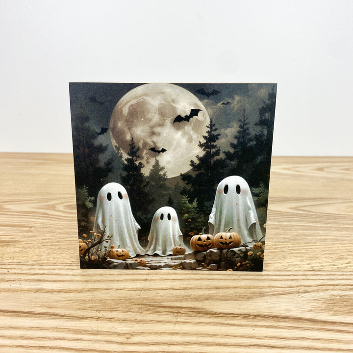 Halloween-themed card with ghosts, pumpkins, and a full moon on a wooden surface.